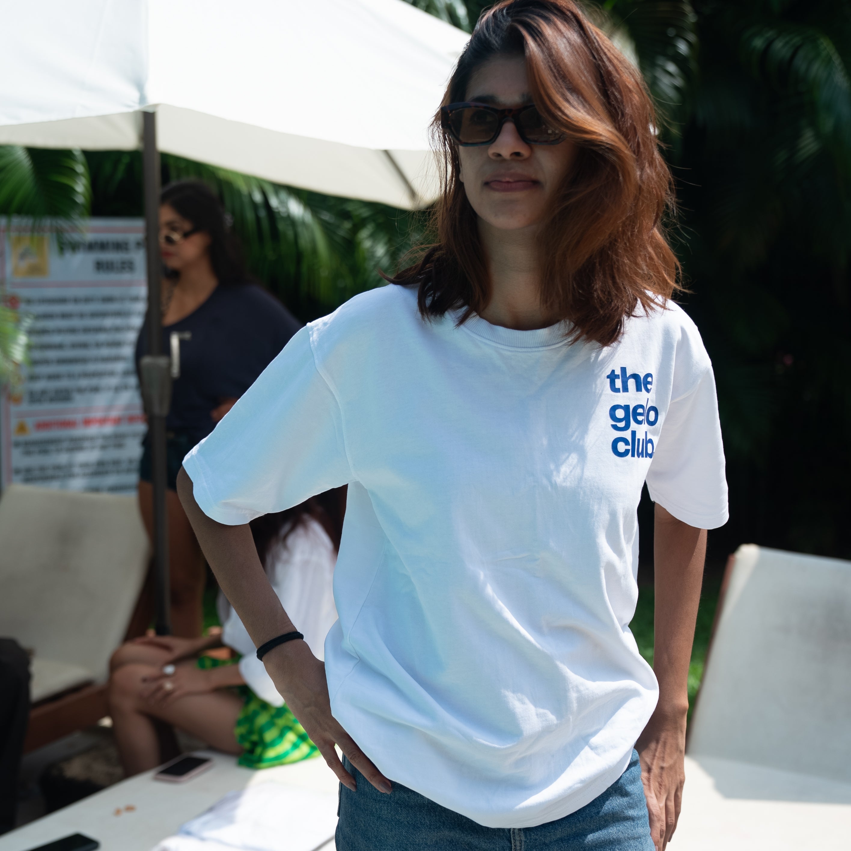 Person wearing Gelo white t-shirt, standing outdoors near the pool under an umbrella.